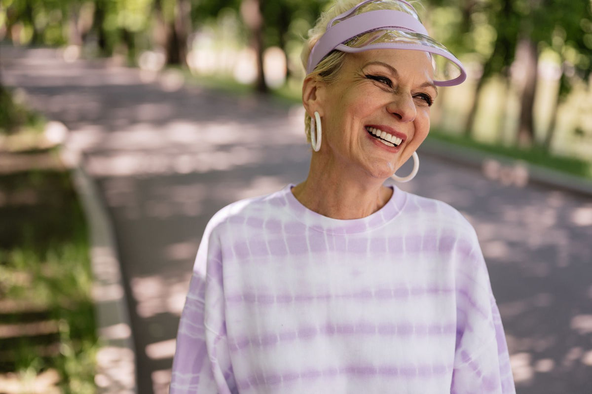 An older woman laughing on a sunny park path in a lavender sun visor
